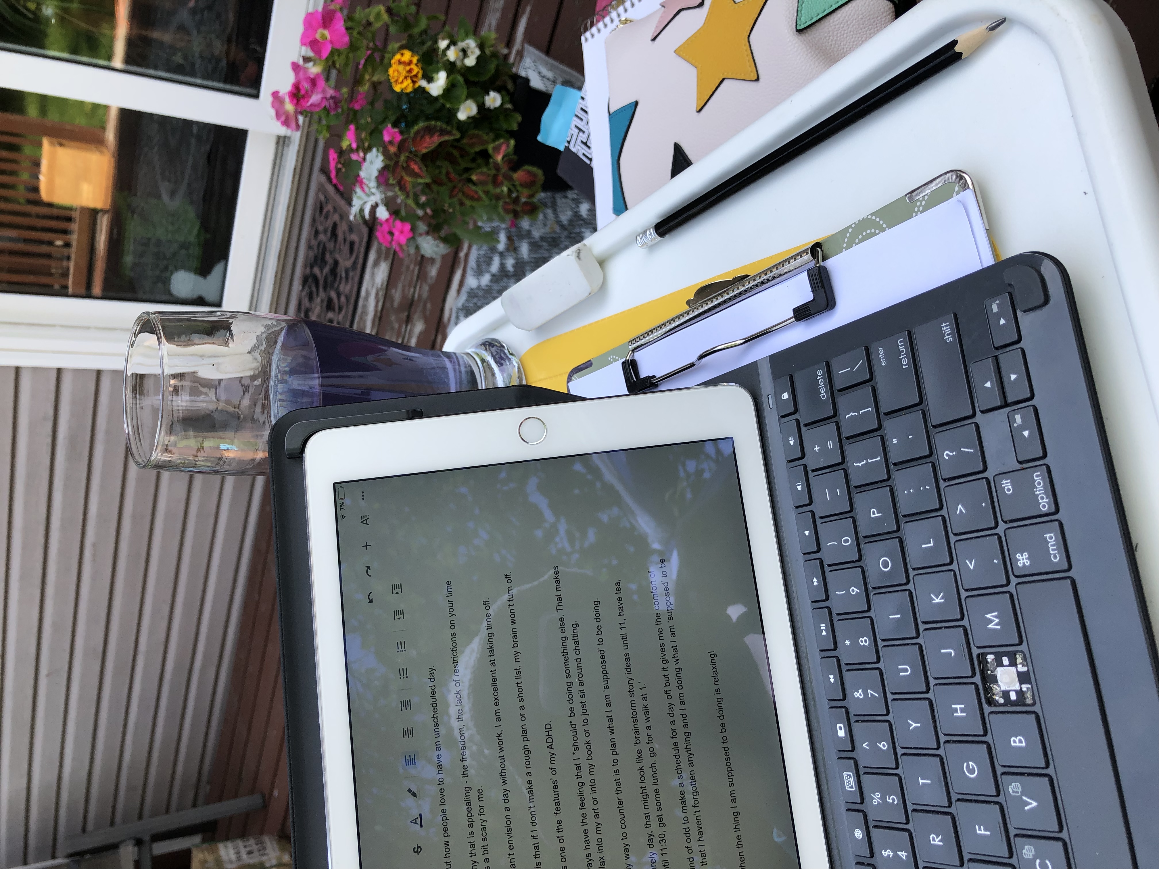 The author’s iPad sits on a white table on a patio, there is a tall glass of blue tea in the background and there are notebooks and a pencil case nearby. There is a pot of flowers on the patio behind the table.