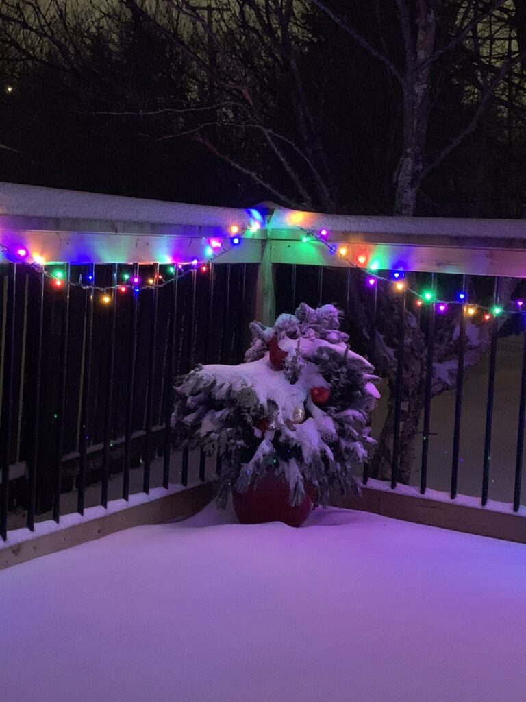 Photo of light snow on the corner of my patio where I have a decorative arrangement of festive greenery in a flowerpot and there’s a  string of Christmas lights on the railing above it.