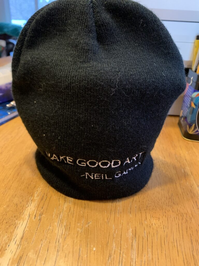 A black beanie with white text that reads ‘Make Good Art’ - Neil Gaiman. It appears to be standing on its own on my wooden table but it is actually serving as a tea cozy for my small teapot.?